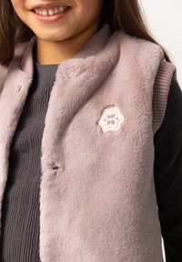 Jasnoróżowa kamizelka z futra syntetycznego z prążkowanym kołnierzem, z kremową gwiazdkową naszywką z napisem "NP PS" na środku.