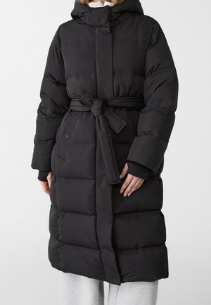 Femme portant un long manteau matelassé noir avec une capuche et une ceinture nouée, debout devant un fond blanc uni.