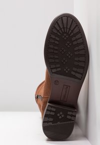 Botte en daim marron avec semelle noire texturée visible, motif de semelle et fermeture éclair latérale, posée contre un mur d’angle blanc.