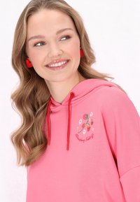 Felpa con cappuccio rosa, con lacci rossi e ricami decorativi che raffigurano fragole e fiori. Tessuto morbido, vestibilità rilassata.