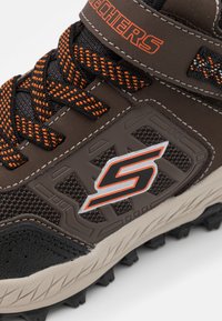 Zapatilla deportiva marrón con parte superior de tela texturizada, acentos naranjas, cordones negros, suela de goma y detalle del logo en el lateral.