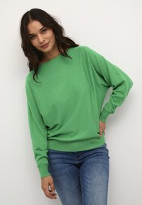 Groene, lichte trui met een losse pasvorm, geribbelde manchetten en zoom. Gecombineerd met blauwe spijkerbroek, tegen een neutrale achtergrond.