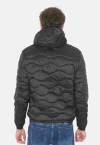 Giacca imbottita nera con cappuccio, caratterizzata da una texture trapuntata e una finitura esterna liscia. La parte posteriore presenta un orlo arrotondato e polsini elasticizzati.