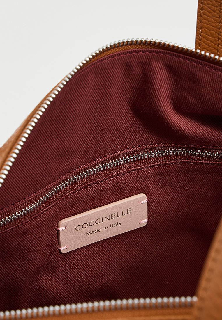 Brązowa skórzana torba z bordowym wnętrzem, z metalowym suwakiem i różową metką z napisem "Coccinelle Made in Italy".