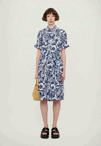 Vestido azul com padrão floral, com mangas curtas em balão, cintura franzida e saia até aos joelhos; combinado com uma bolsa de palha e sandálias grossas.