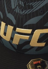Juodas kepurė su tekstūruotu auksiniu "UFC" logotipu, sudėtingu juodu ir teal spalvos raštu, bei šešiakampe juoda ir auksinė etikete.