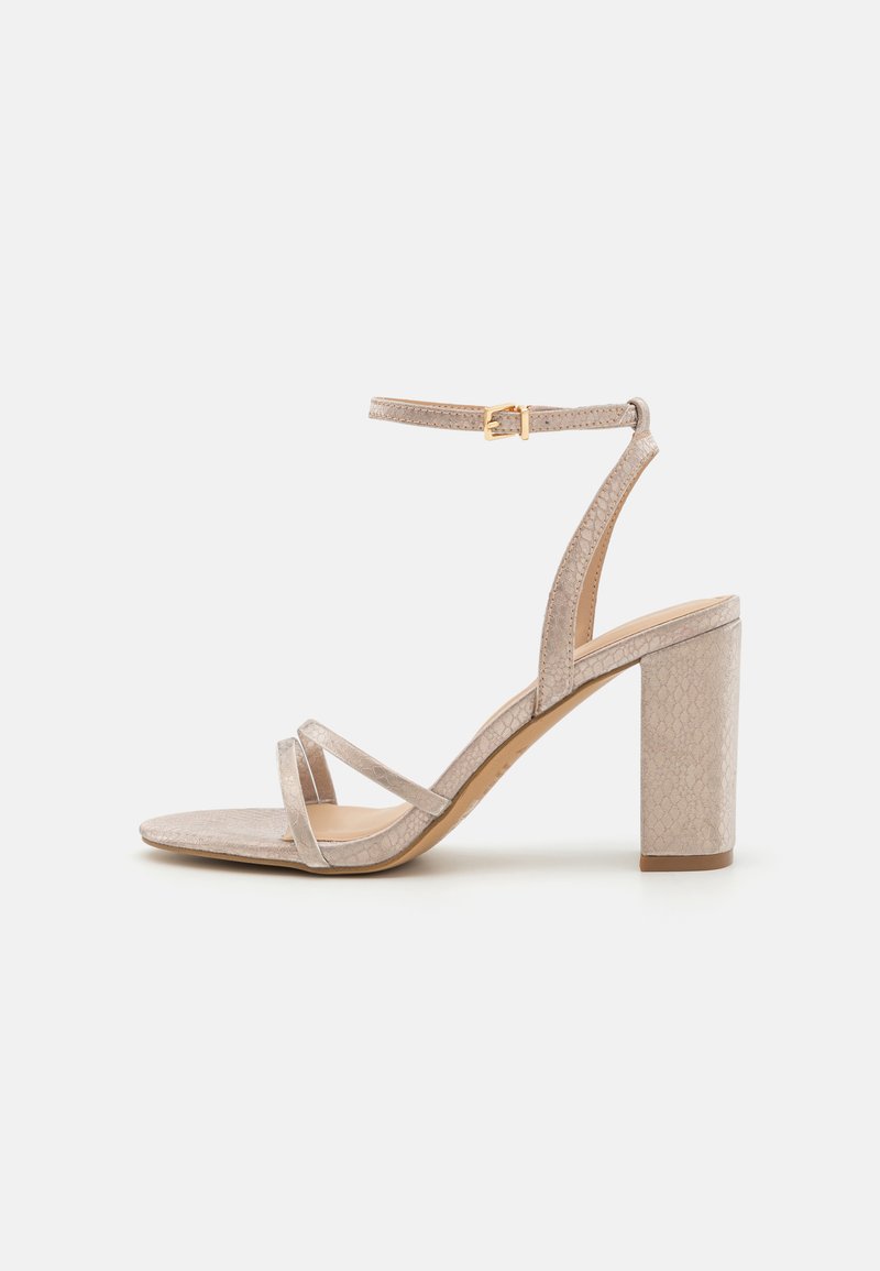 Sandale à talons beige avec une finition texturée, dotée de double brides sur les orteils et d'une bride de cheville ajustable. Design de talon carré.
