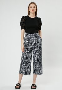 Zwart top met kanten pofmouwen, gecombineerd met wijde broek met een zwart-wit abstract patroon. Zwarte slip-ons maken de look compleet.