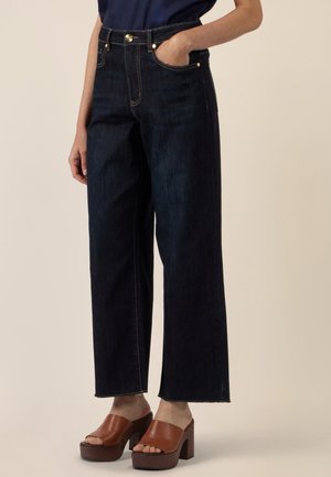 Person trägt hoch taillierte dunkelblaue, weit geschnittene Jeans und braune Plateau-Sandalen mit offenem Zehenbereich und Absatz, eine Hand in der Tasche.