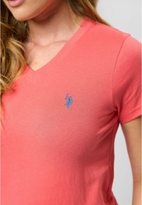 Koralen vrhnji T-shirt iz mehkega bombaža, ki ima majhen modri logotip izšit na levem prsnem delu. Kratke rokave, gladka tekstura.