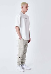 Un giovane con i capelli biondi corti è in piedi di lato, indossando una t-shirt bianca oversize, pantaloni cargo chiari, sneaker bianche e un bracciale d'argento.