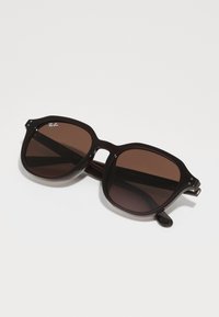 Lunettes de soleil surdimensionnées teintées de brun avec une monture noire épaisse. Les caractéristiques incluent des bords arrondis, des accents en métal et un logo sur le coin supérieur gauche de la lentille.