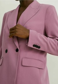 Personne ajustant le revers d'un blazer lavande croisé à boutons noirs et poches à rabat.