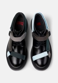 Zapatos negros con suelas de goma anchas y correas elásticas en tonos azul verdoso, gris y azul claro. Presentan una punta redondeada y una plantilla acolchada.
