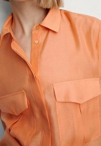 Camicia button-up color pesca realizzata in tessuto leggero, con colletto, due tasche frontali e una vestibilità rilassata. Texture liscia.