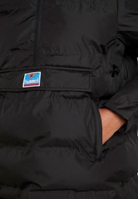 Sort puffjakke med lynlås, der har en frontlomme og et fremtrædende logo-patch i blå og rød. Glat, shiny stoftekstur.
