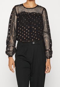 Blusa nera trasparente con scollatura arricciata, maniche lunghe a sbuffo e motivo a pois color rame. Abbinata a pantaloni neri a vita alta.
