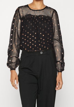 Blusa nera trasparente con scollatura arricciata, maniche lunghe a sbuffo e motivo a pois color rame. Abbinata a pantaloni neri a vita alta.