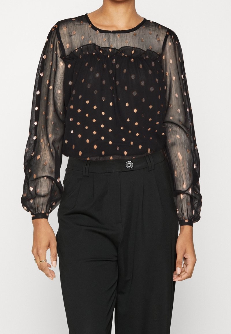 Blusa nera trasparente con scollatura arricciata, maniche lunghe a sbuffo e motivo a pois color rame. Abbinata a pantaloni neri a vita alta.