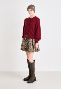 Cardigan en laine rouge mélangé avec des boutons, associé à une jupe à motifs léopard. Le modèle porte des bottes marron au genou et un bonnet marron côtelé.