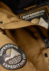 Brązowa ocieplana kurtka z czarną łatą z górskim wzorem i napisem logo. Gładki materiał, zamek błyskawiczny i napy.