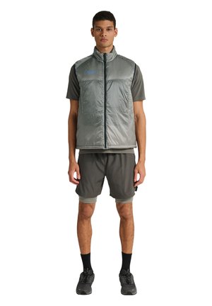 Jonge man met een grijze, mouwloze gewatteerde bodywarmer over een grijs T-shirt met korte mouwen, grijze korte broek, zwarte sokken en zwarte sneakers, staand en recht vooruit kijkend.