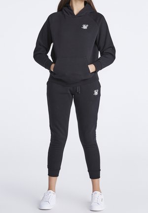 Femme portant un sweat à capuche noir et un jogging avec des logos blancs, debout les mains dans la poche du sweat, chaussée de baskets blanches.