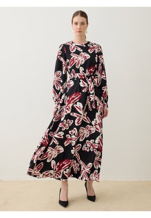 Donna che indossa un lungo vestito nero con grandi stampe floreali rosse e bianche, maniche lunghe, cintura in vita e tacchi alti neri, in piedi contro un muro bianco.