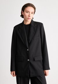 Sacou negru cu un finisaj ușor texturat, având două buzunare frontale și nasturi decorativi. Formă ajustată cu rever înclinat.