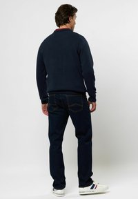 Mann steht mit dem Rücken zur Kamera, trägt einen marineblauen Pullover, dunkelblaue Jeans und weiße Sneaker mit roten und blauen Streifen.
