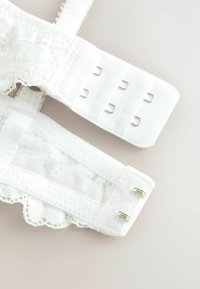Reggiseno bianco in pizzo con fascia caratterizzata da un bordo in pizzo testurizzato, ganci e chiusure a occhiello. Tessuto liscio con dettagli delicati e spalline regolabili.