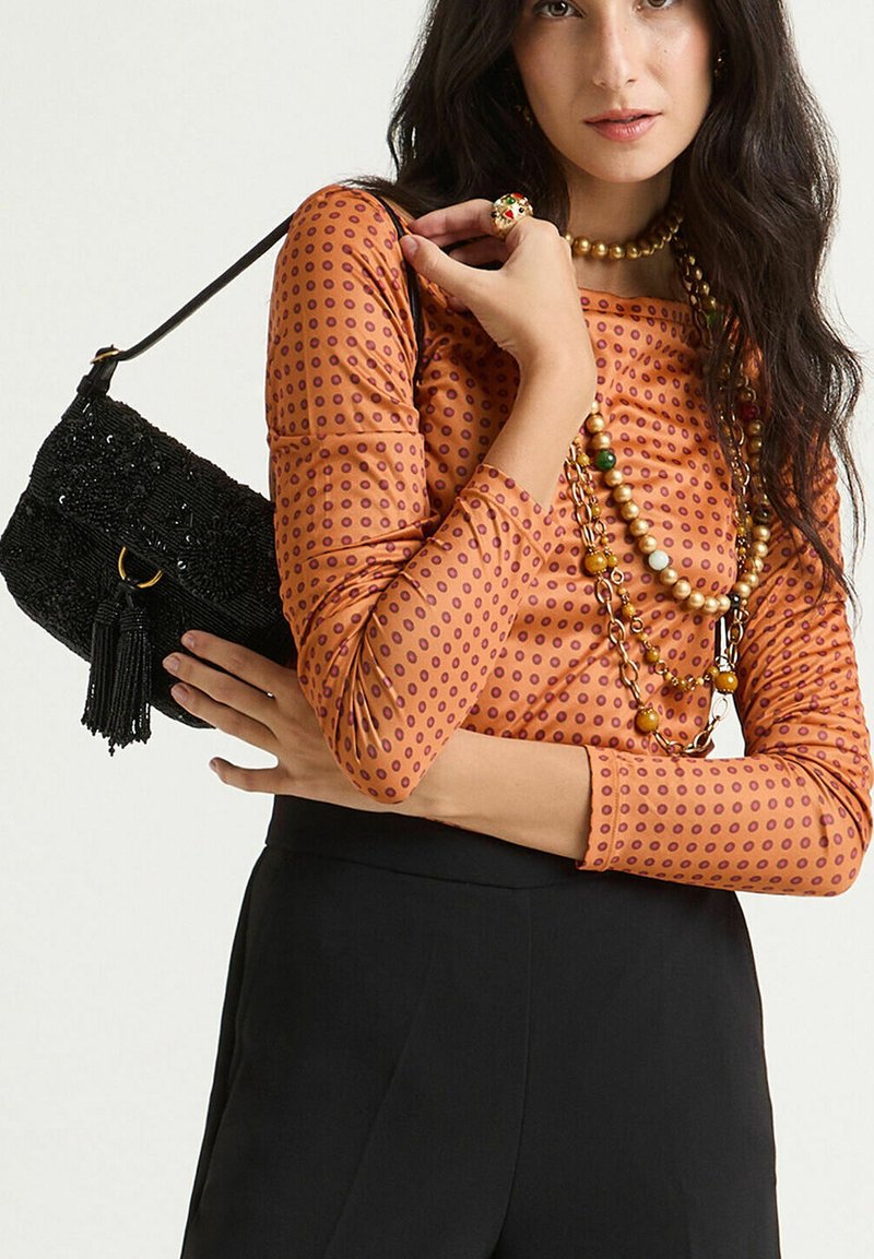 Borsa a tracolla nera con perline e dettaglio a nappina; indossata con una maglia arancione con pois viola e collane a strati. Pantaloni neri.