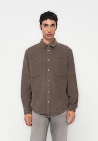 Chemise en velours côtelé marron avec boutons à l'avant, manches longues et deux poches poitrine. Elle présente un col classique et un tissu texturé.