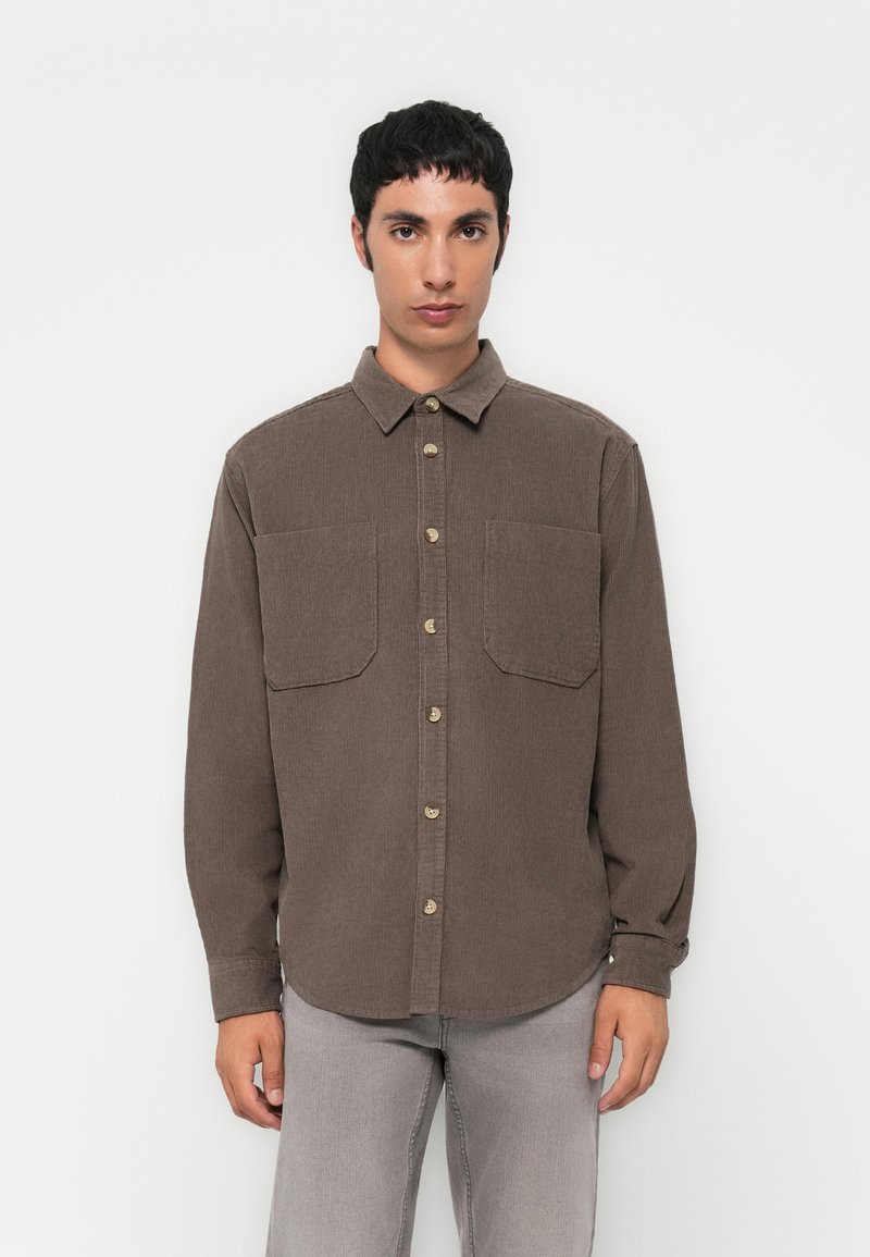 Chemise en velours côtelé marron avec boutons à l'avant, manches longues et deux poches poitrine. Elle présente un col classique et un tissu texturé.