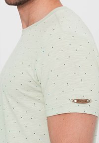 Jasnozielona koszulka z krótkim rękawem, pokryta nadrukiem liter w kolorze czarnym i niebieskim. Posiada brązową metkę z logo na lewym rękawie.