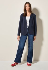 Blazer bleu marine à coupe décontractée, deux poches avant, associé à un jean bleu à jambes larges et des baskets rouges avec des rayures blanches.