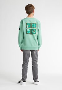 Sweatshirt vert menthe avec un graphique au dos en vert et turquoise, présentant un design rectangulaire. Porté avec un jean gris et des chaussures blanches.