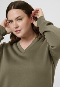 Femme aux cheveux châtain mi-longs portant un pull en V vert olive, ajustant ses cheveux derrière ses oreilles devant un fond uni.