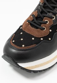 Sneaker nera e marrone con superficie testurizzata, decorata con borchie dorate e una suola intermedia a strisce bianche e dorate. Punta rotonda e lacci neri.