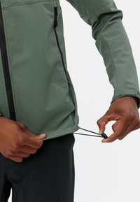 Grüne Softshelljacke mit einem Frontreißverschluss, verstellbarem Saum und strukturiertem Stoff. Der Träger zieht an einem Kordelzug am unteren Ende für die Passformanpassung.