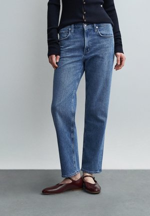 Femme portant un jean bleu droit, un pull à côtes bleu marine foncé et des chaussures plates rouge foncé avec brides à la cheville, debout devant un fond gris.