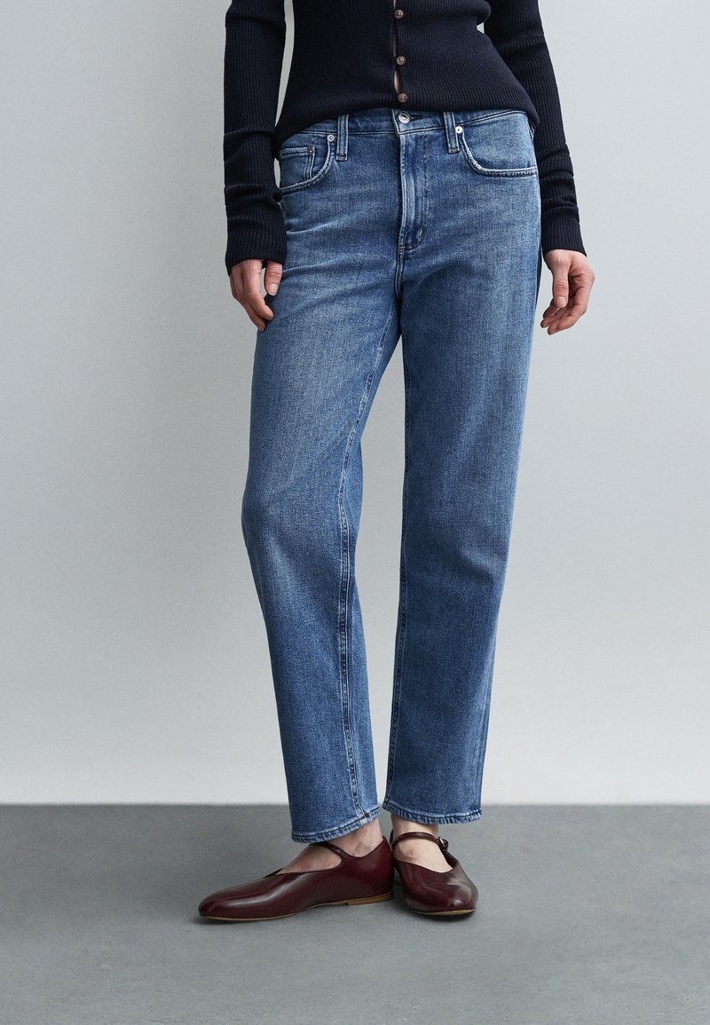 Γυναίκα που φοράει ίσια μπλε τζιν, σκούρο μπλε πουλόβερ με ριπ υφή και σκούρα κόκκινα επίπεδα παπούτσια με λουράκια στον αστράγαλο, στέκεται μπροστά σε γκρι φόντο.