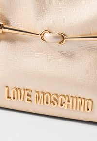 Zlatá metalická kabelka s textúrovaným povrchom, so reťazkou a embosovaným názvom značky "LOVE MOSCHINO" v zlatých písmenách.