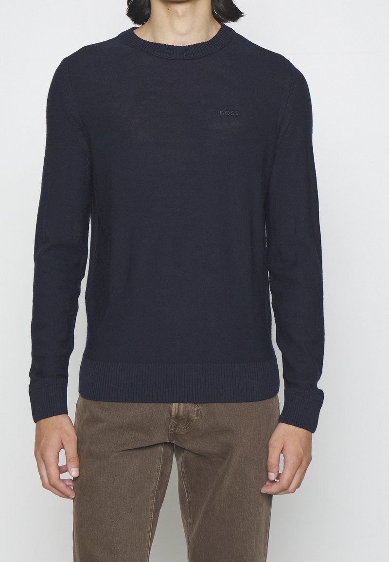 Homme portant un pull col rond bleu marine uni et un pantalon marron, debout devant un fond blanc.