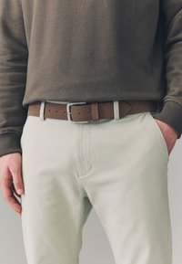 Brown fabric belt with silver rectangular buckle, worn over light-colored pants and a dark sweatshirt with ribbed cuffs.
