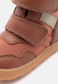 La scarpa in pelle marrone presenta una superficie texturizzata, doppie fibbie in velcro, un design a righe in una tonalità più chiara e una suola in gomma per un migliore grip.