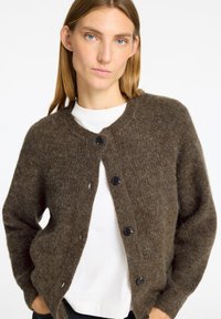 Frau mit glattem, hellbraunem Haar, die eine braune, flauschige, geknöpfte Strickjacke über einem weißen Shirt trägt und mit den Händen in den Taschen posiert.