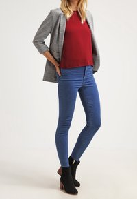 Blazer grigio sopra una maglietta bordeaux, abbinato a jeans skinny blu e stivaletti neri con tacco basso a blocco.