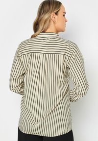 Camisa de botón beige con rayas verticales negras, de manga larga, con detalle de yugo y corte relajado. Textura suave, tejido ligero.