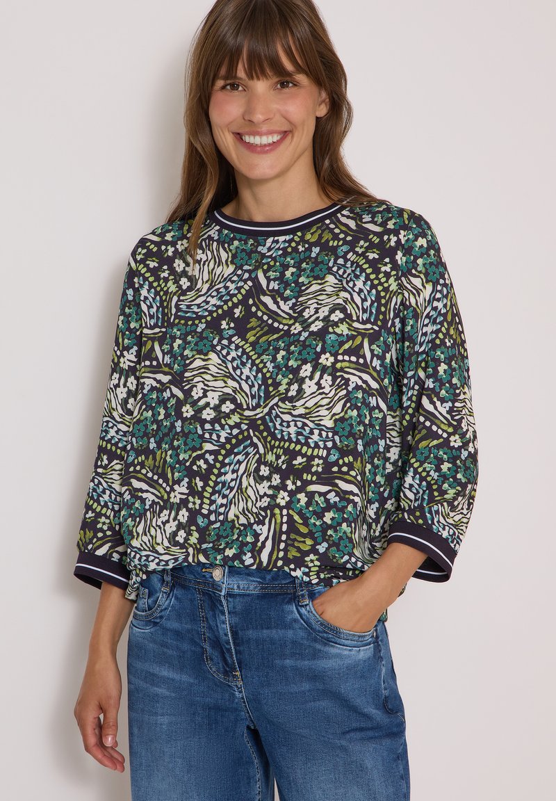 Femme souriante portant un haut à manches longues à motifs floraux et abstraits avec des poignets bleu marine et un jean bleu, debout avec une main dans la poche.